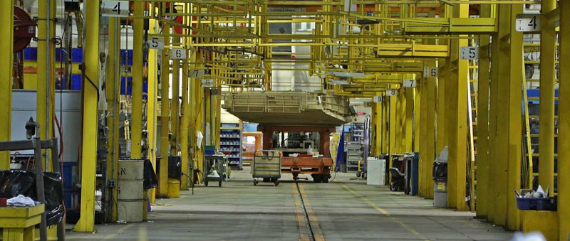 A factory aisle lined with yellow hoists, with a vehicle hull moving overhead.