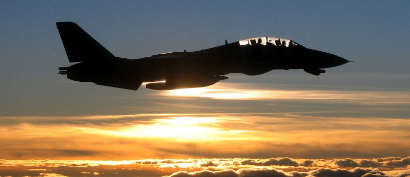 A fighter jet in silhouette flies above clouds at sunset.