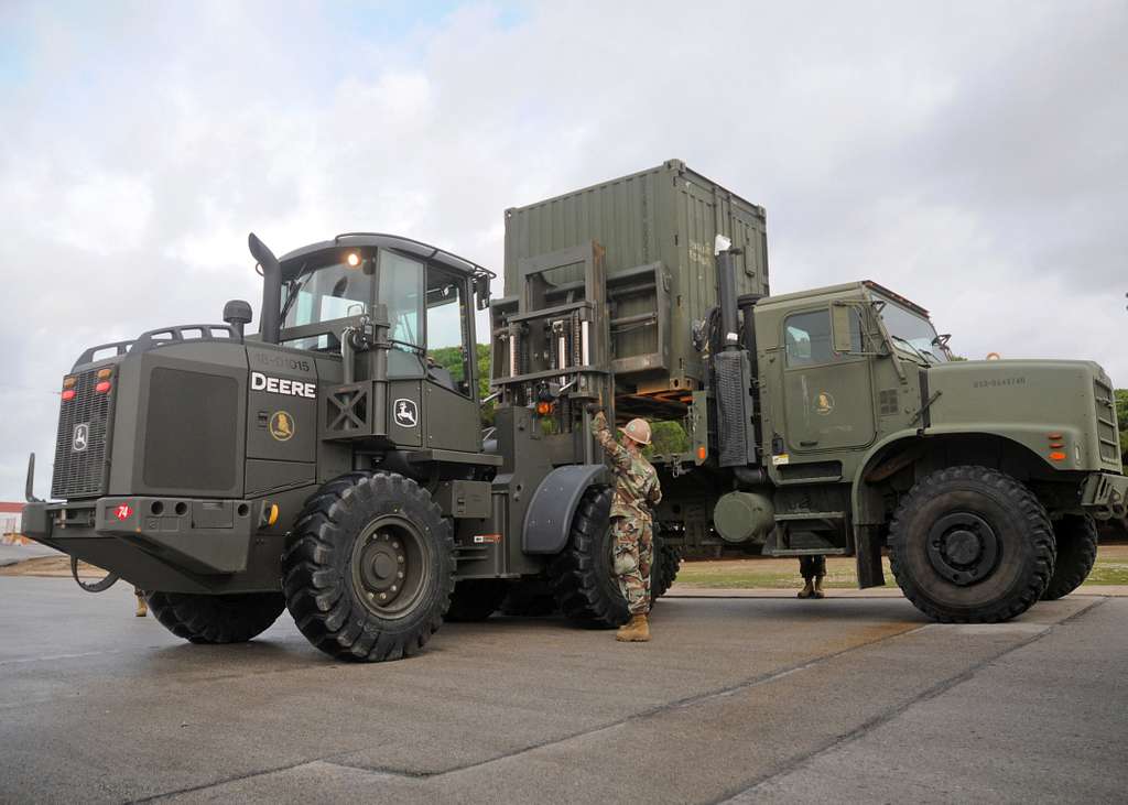 A service member guides a shipping container onto the bed of a military truck.